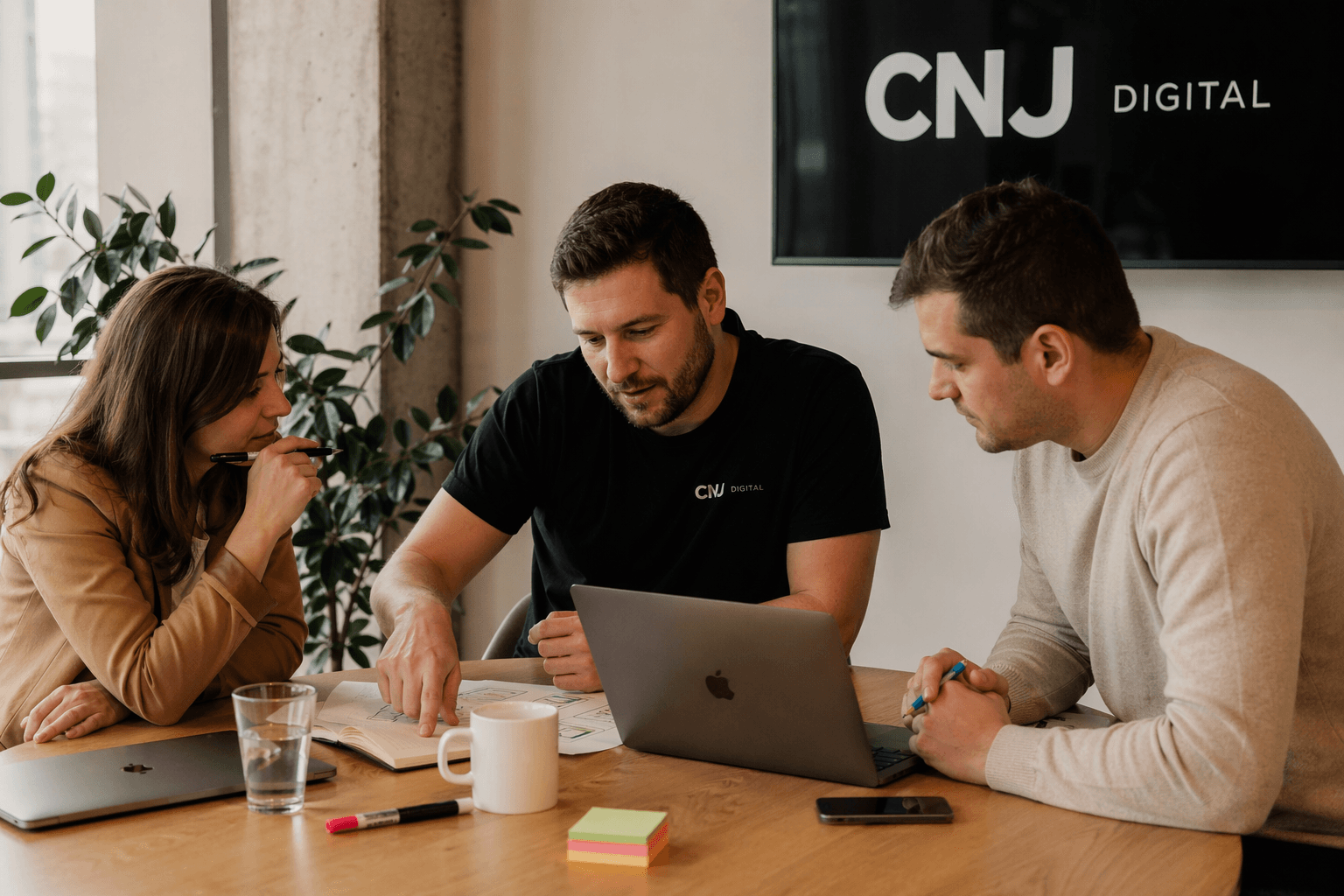 Three colleagues at CNJ Digital collaborate at a meeting table, reviewing documents and laptops. The workspace includes stationery, a glass of water, and a coffee mug, with a CNJ Digital logo visible on a screen in the background.